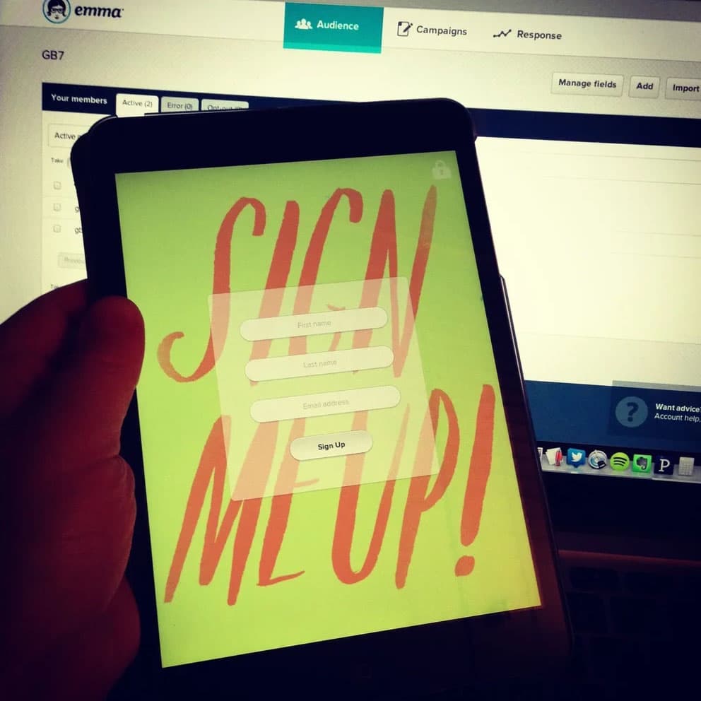 Guestbook running on an iPad Mini held in front of an Emma desktop screen, showing a 'SIGN ME UP!' email signup form.